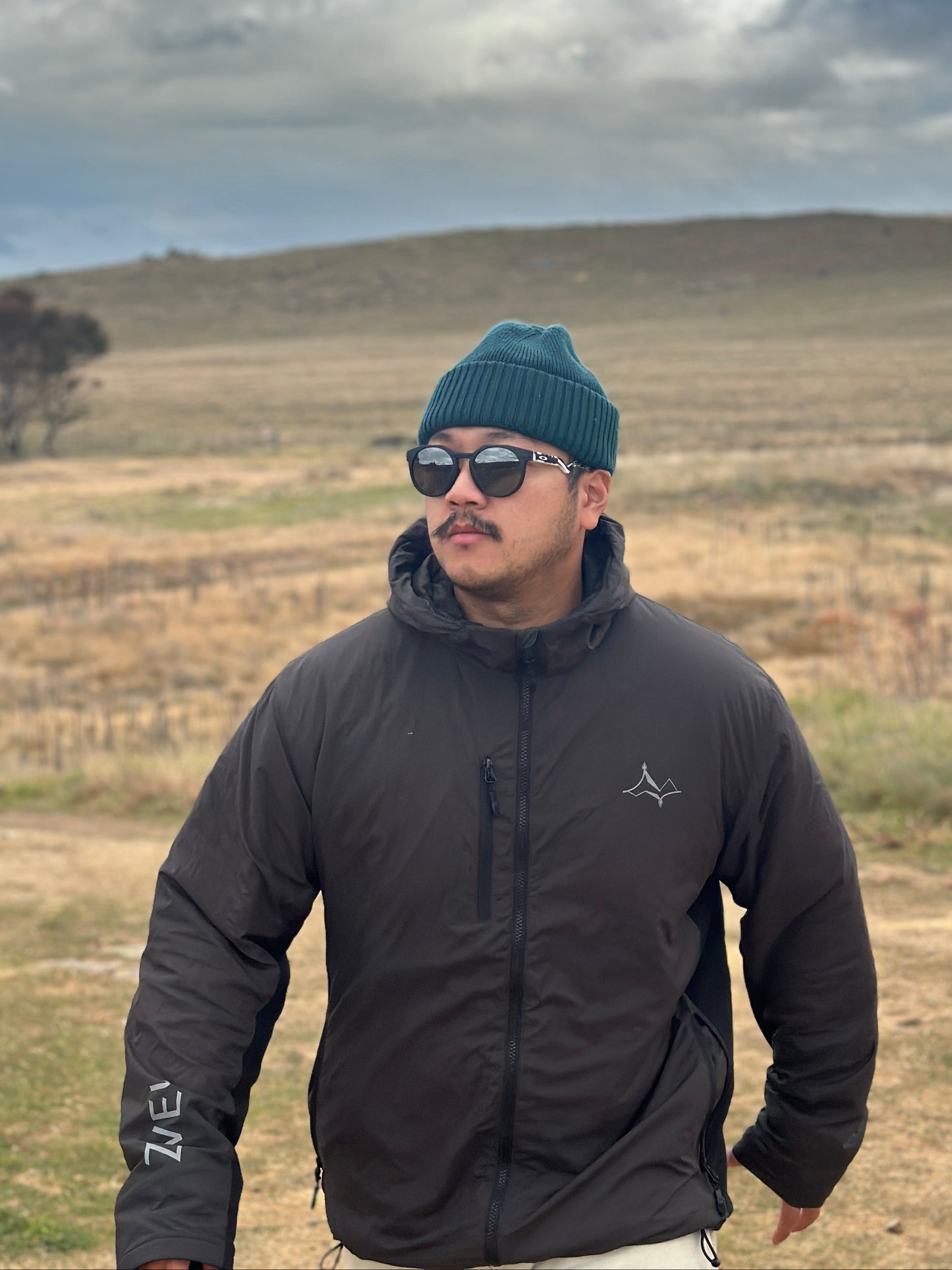 Man wearing a black jacket and blue beanie standing in a field with a blue sky and clouds.