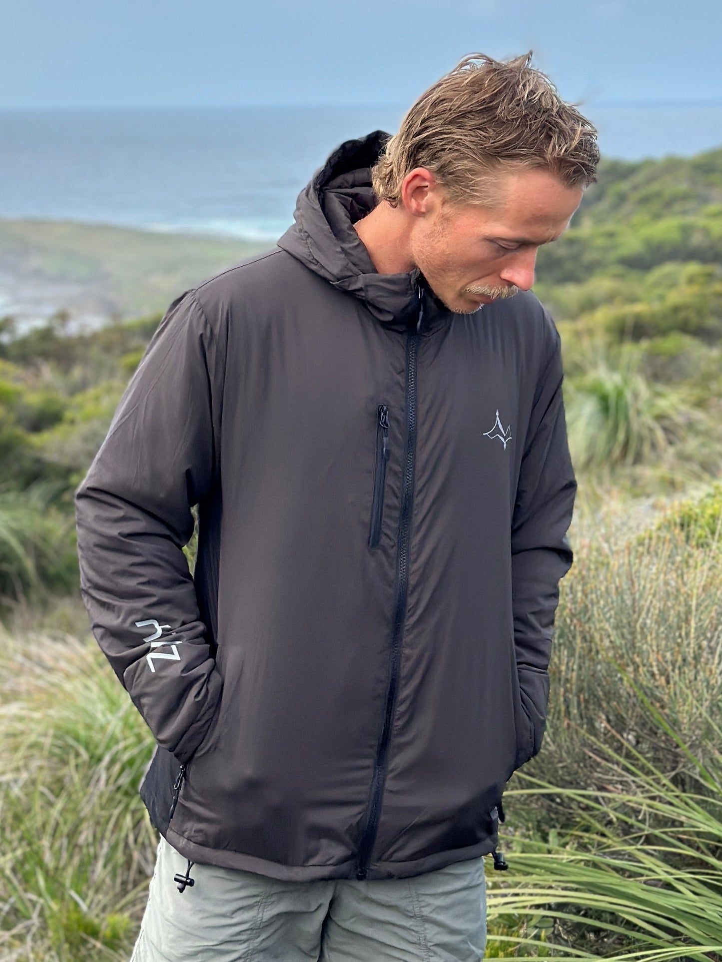 A man wearing a dark gray hooded windbreaker jacket and light gray shorts stands outdoors on a grassy hill overlooking the ocean under a blue sky with clouds.