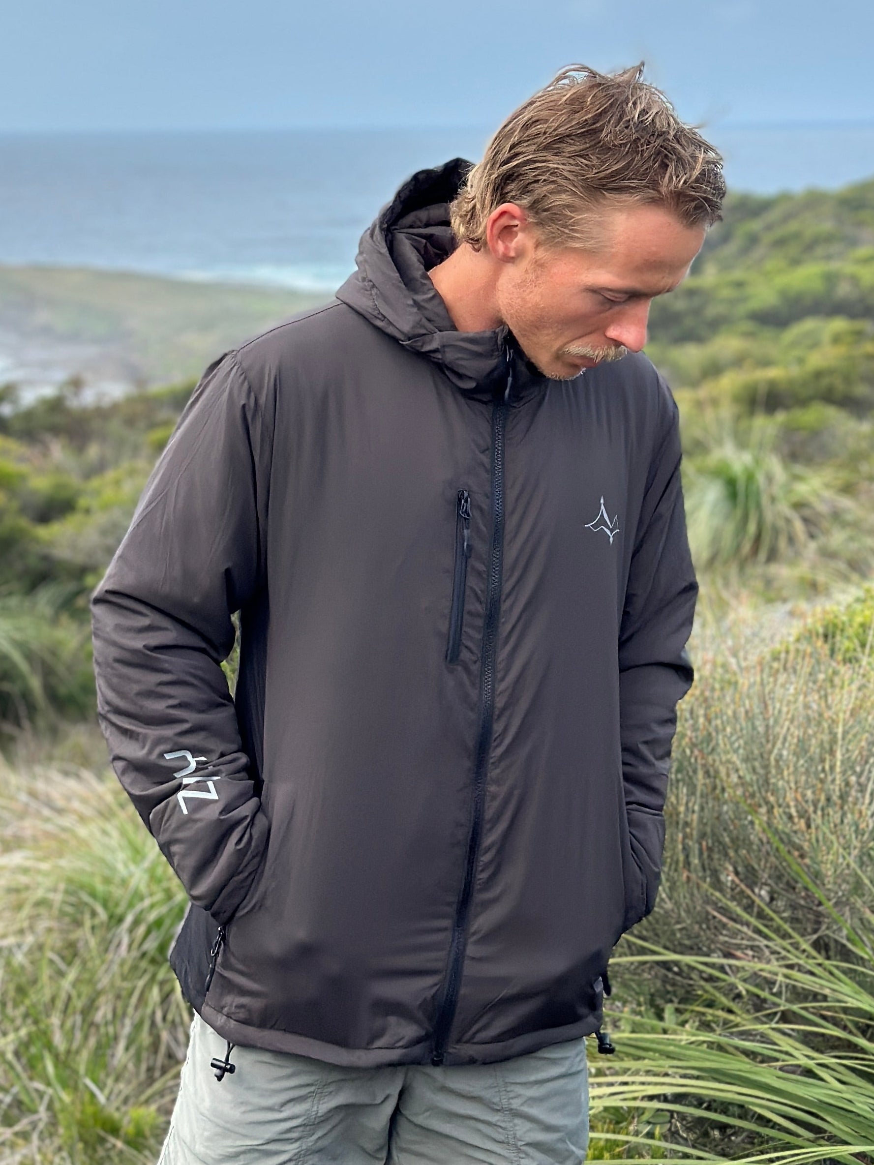 A man wearing a dark gray hooded windbreaker jacket and light gray shorts stands outdoors on a grassy hill overlooking the ocean under a blue sky with clouds.